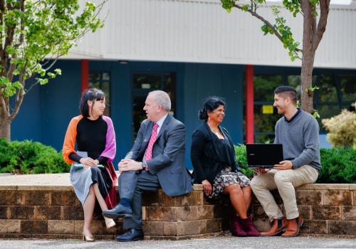 Group of 4 adults sitting outside talking in pairs. 