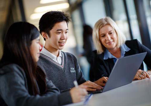 Teacher sitting with two high school students looking at a laptop. 