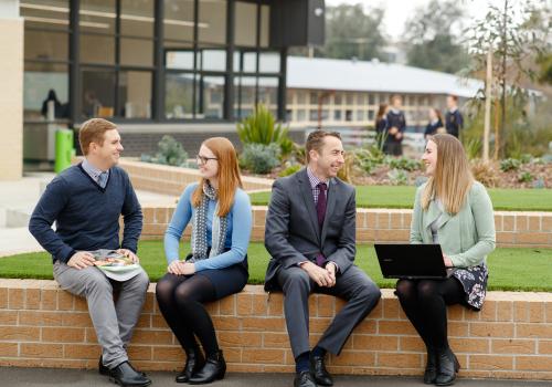 Four adults sitting outside on brick fence talking. 