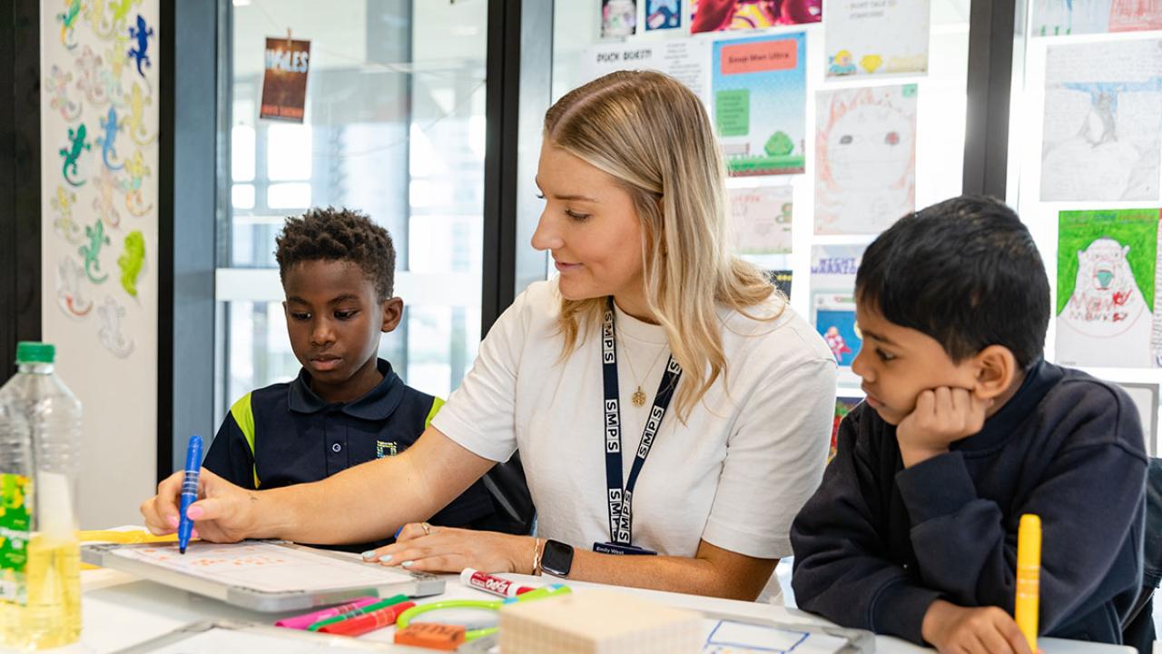 Teacher seated with 2 primary school students at their desk writing on a mini whiteboard