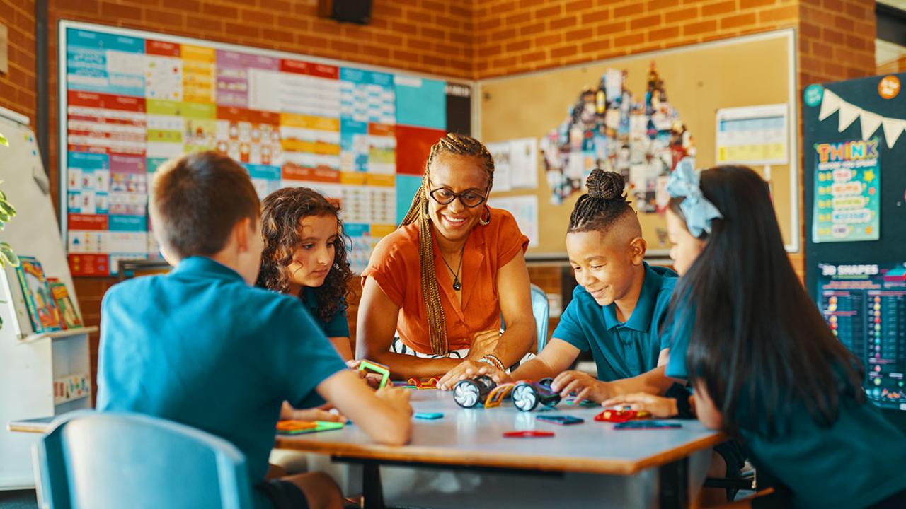 Teacher is seated collaborating with 4 primary school students at desk 