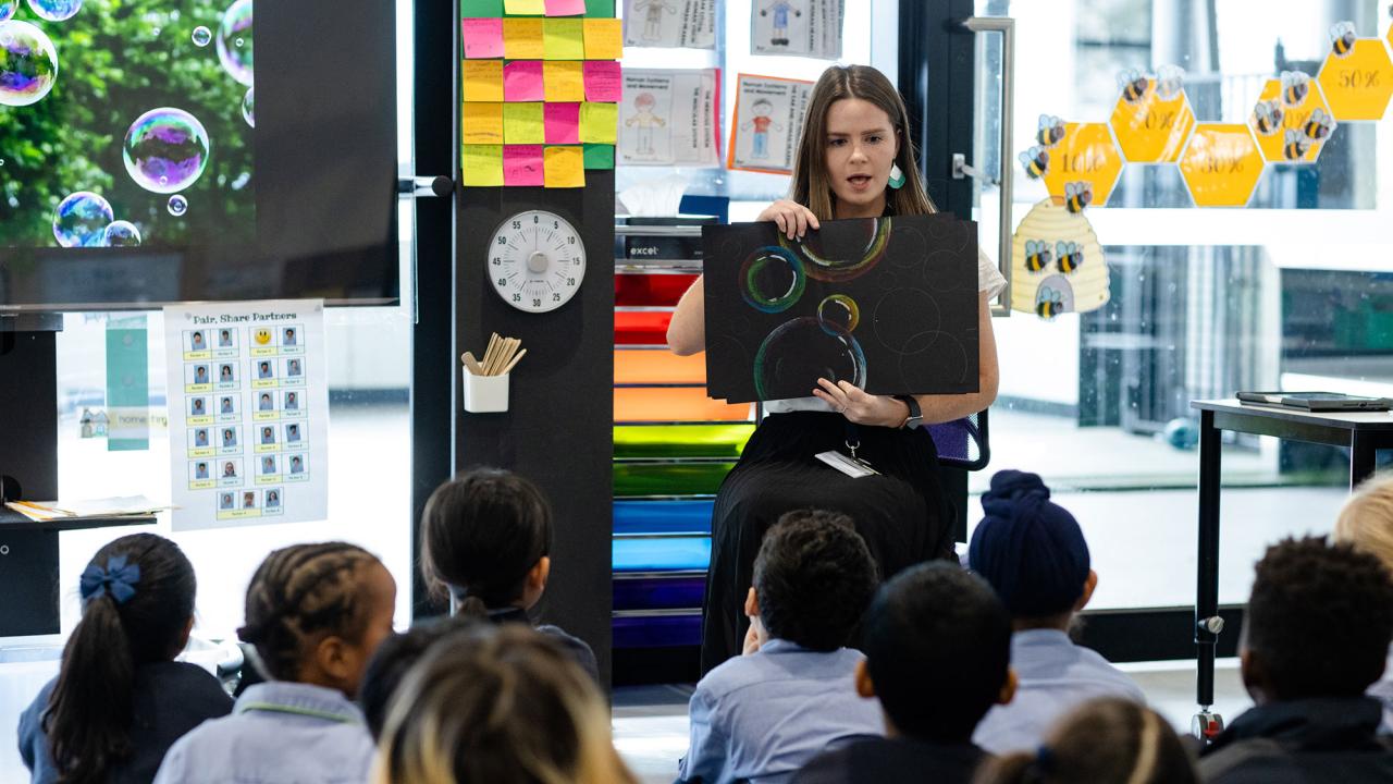 Teacher is seated in front of class of primary school students holding artwok