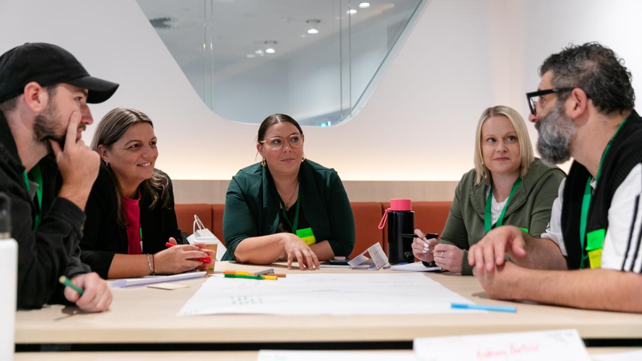 5 participants seated around table in discussion with paper and textas