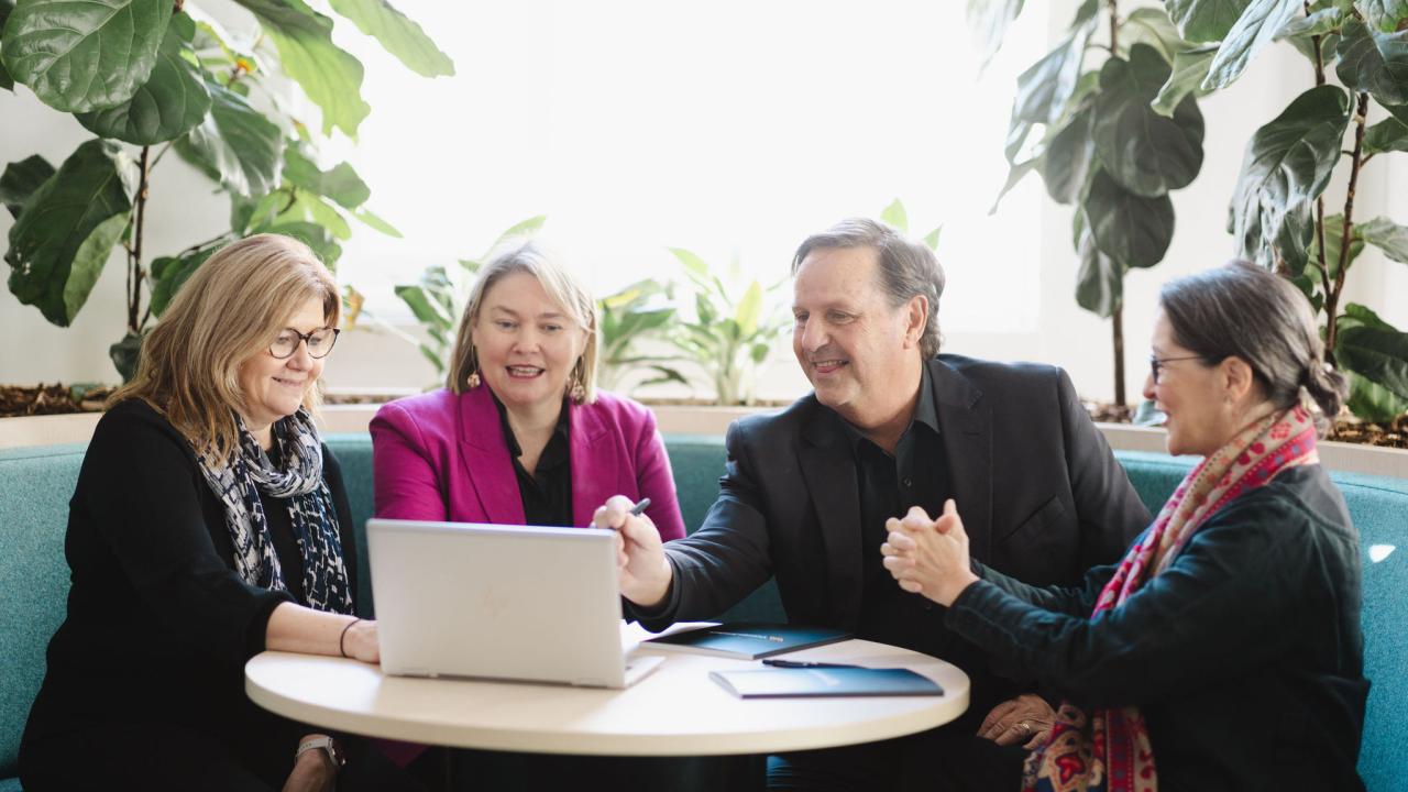 Team of 4 are working together at table focused on a laptop