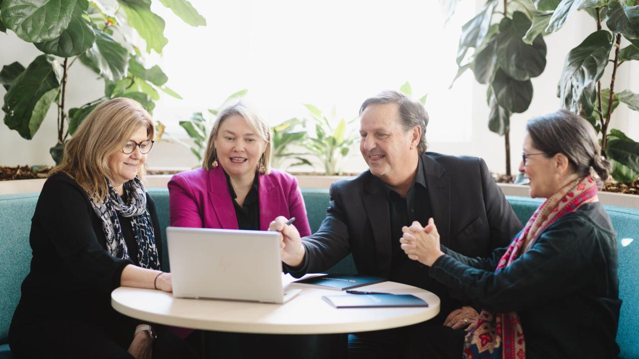 Team of 4 are working together at table focused on a laptop