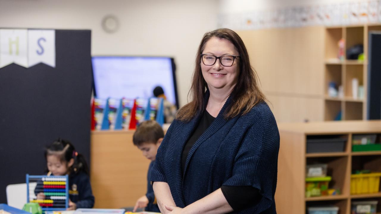 Teacher is standing in the classroom with students seated at tables in the background