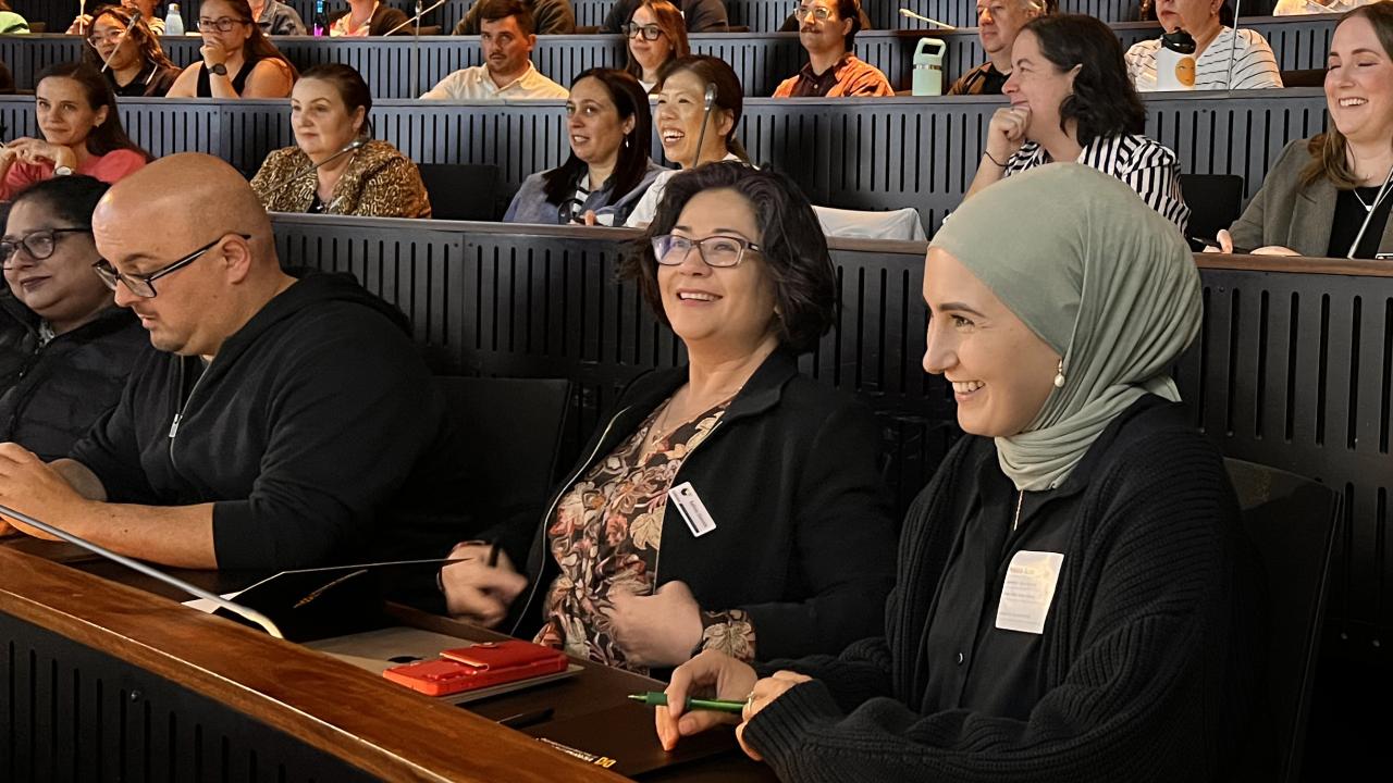 People seated in a modern lecture hall, attentively facing forward, some smiling and engaging with each other.