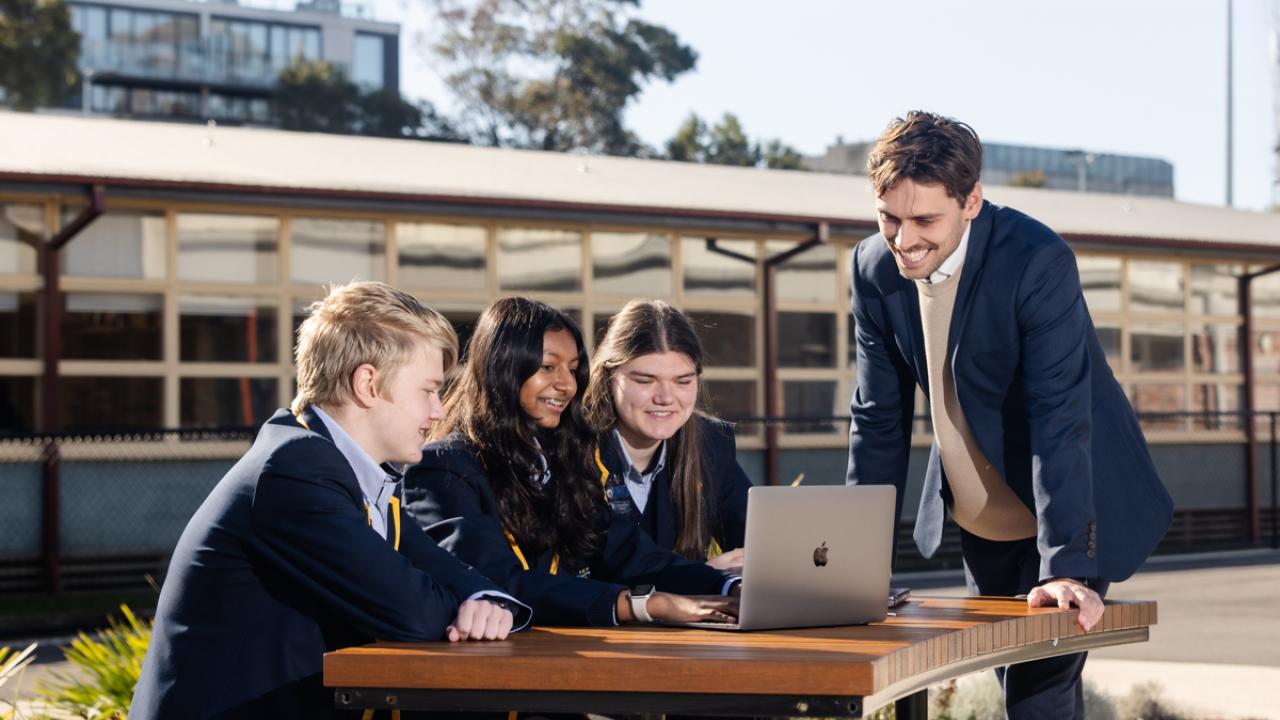 3 high school students are seated at a table with a laptop and a teachers is standing alongside them outside on school grounds