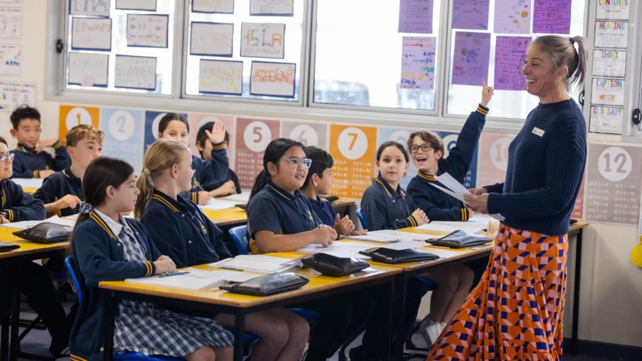 Several students in a class room with one raising their hand to answer a teacher's question. 