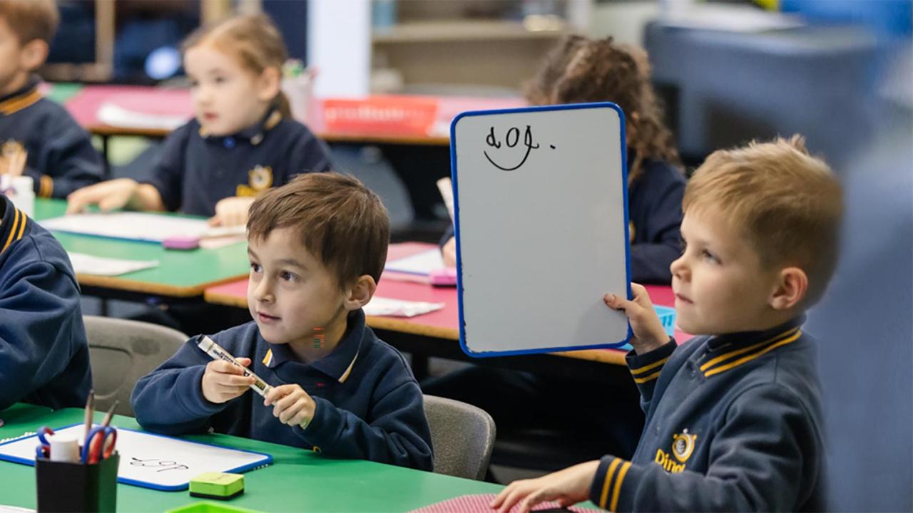 Primary school students at desk writing on small whiteboards. One student is holding up his board with the word dog written on it. This is literacy class