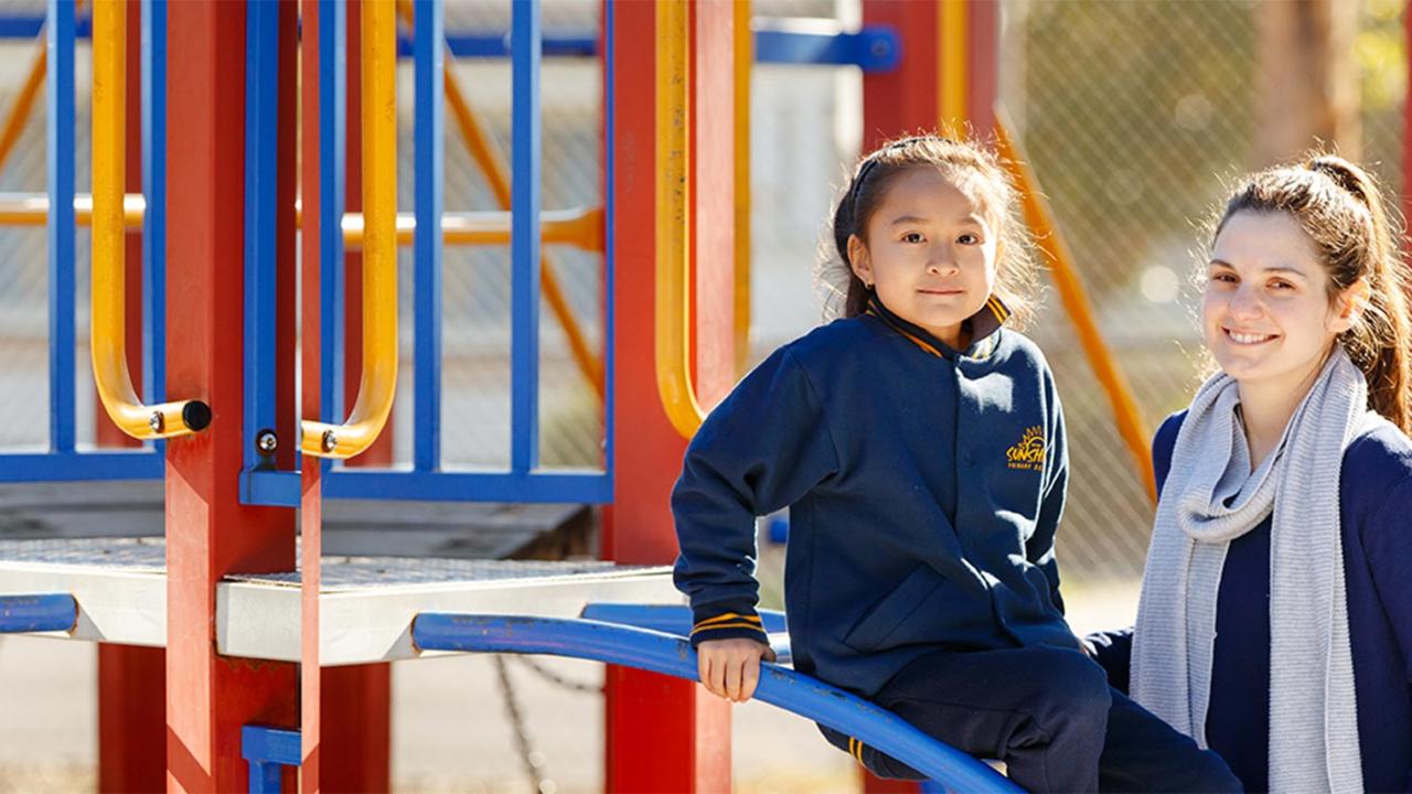 Happy primary school student sitting on slide with female teacher outdoors
