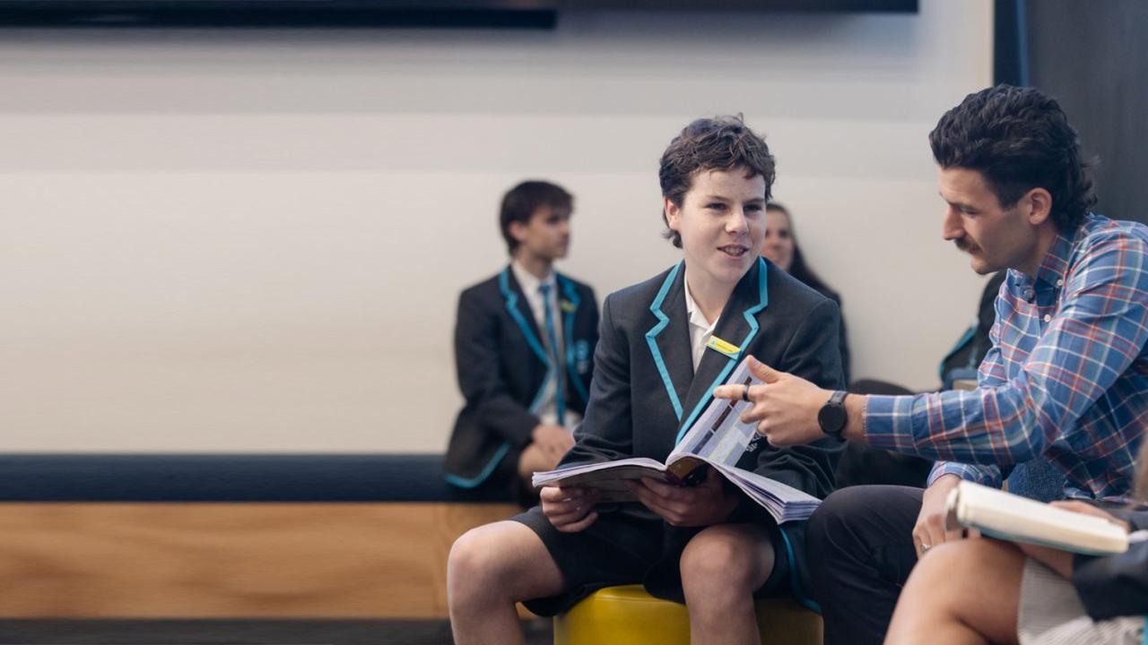 Makle teacher with dark hair and check blue shirt is working through reading material with two secondary students