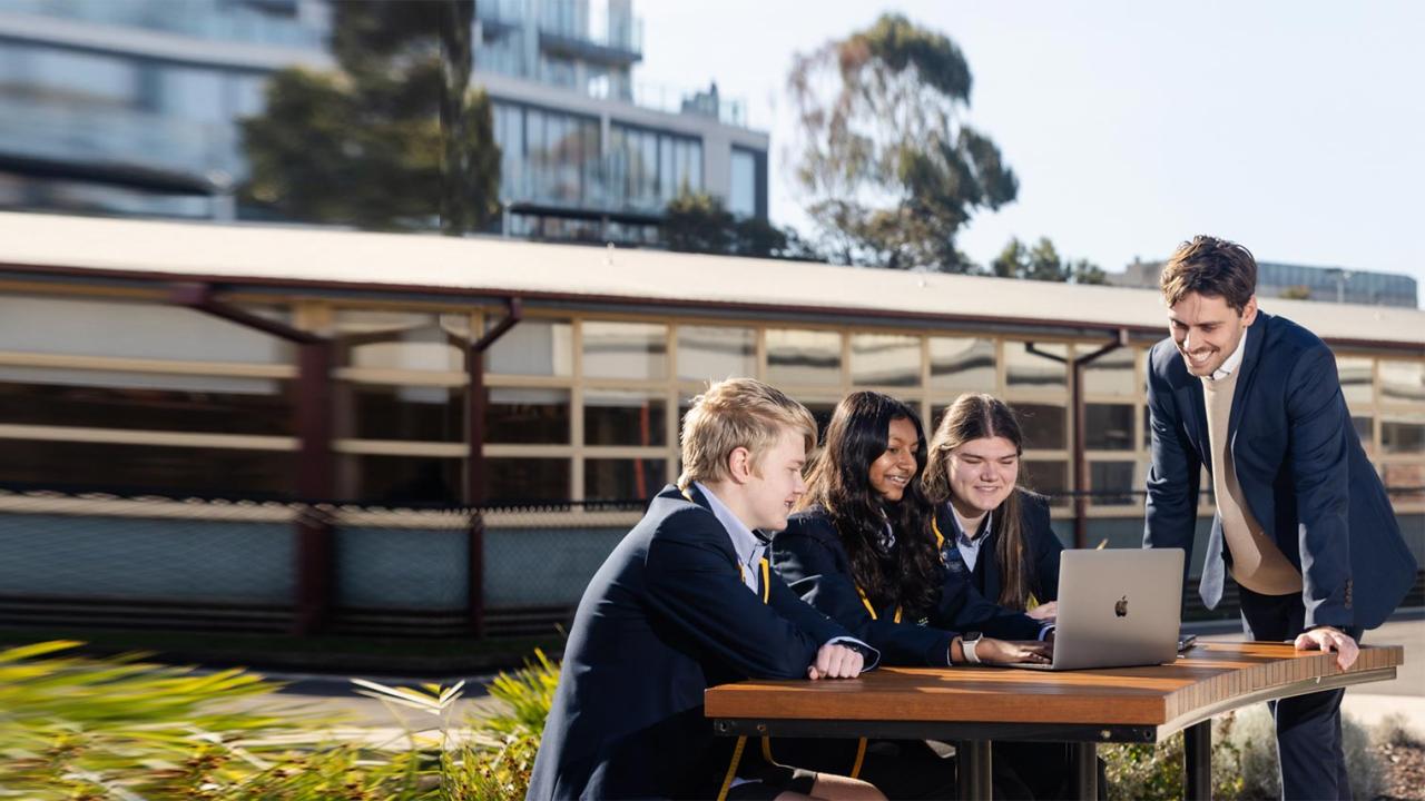 Teacher leading instruction in outdoors while students discuss methods while staring at computer and sitting at outdoor table