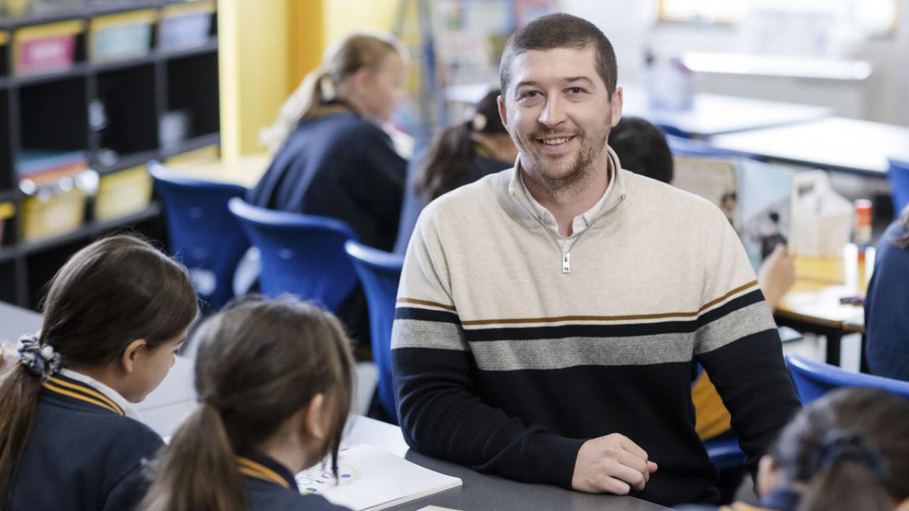 A teacher smiles at the camera from a desk where school children are sitting.