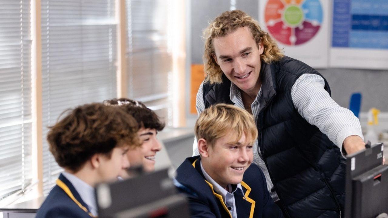 A teacher smiles while pointing to a computer that three children are sitting in front of. ng.
