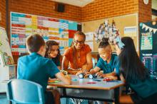 Teacher is seated collaborating with 4 primary school students at desk 