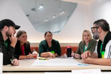 5 participants seated around table in discussion with paper and textas