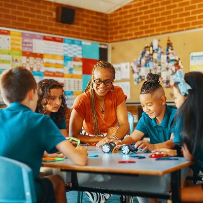 Teacher is seated collaborating with 4 primary school students at desk 