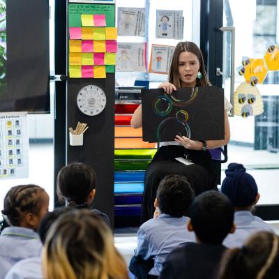 Teacher is seated in front of class of primary school students holding artwok
