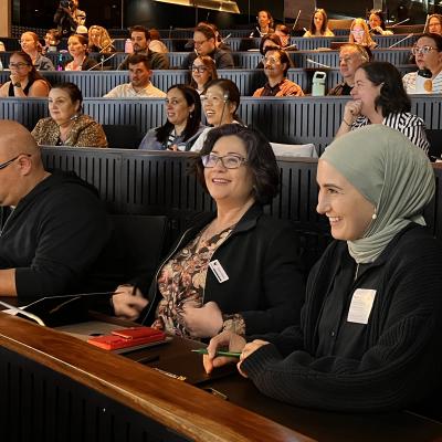 People seated in a modern lecture hall, attentively facing forward, some smiling and engaging with each other.