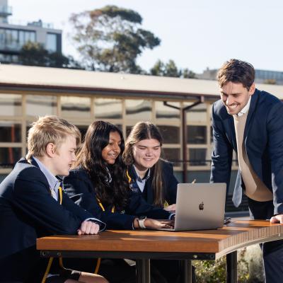 3 high school students are seated at a table with a laptop and a teachers is standing alongside them outside on school grounds