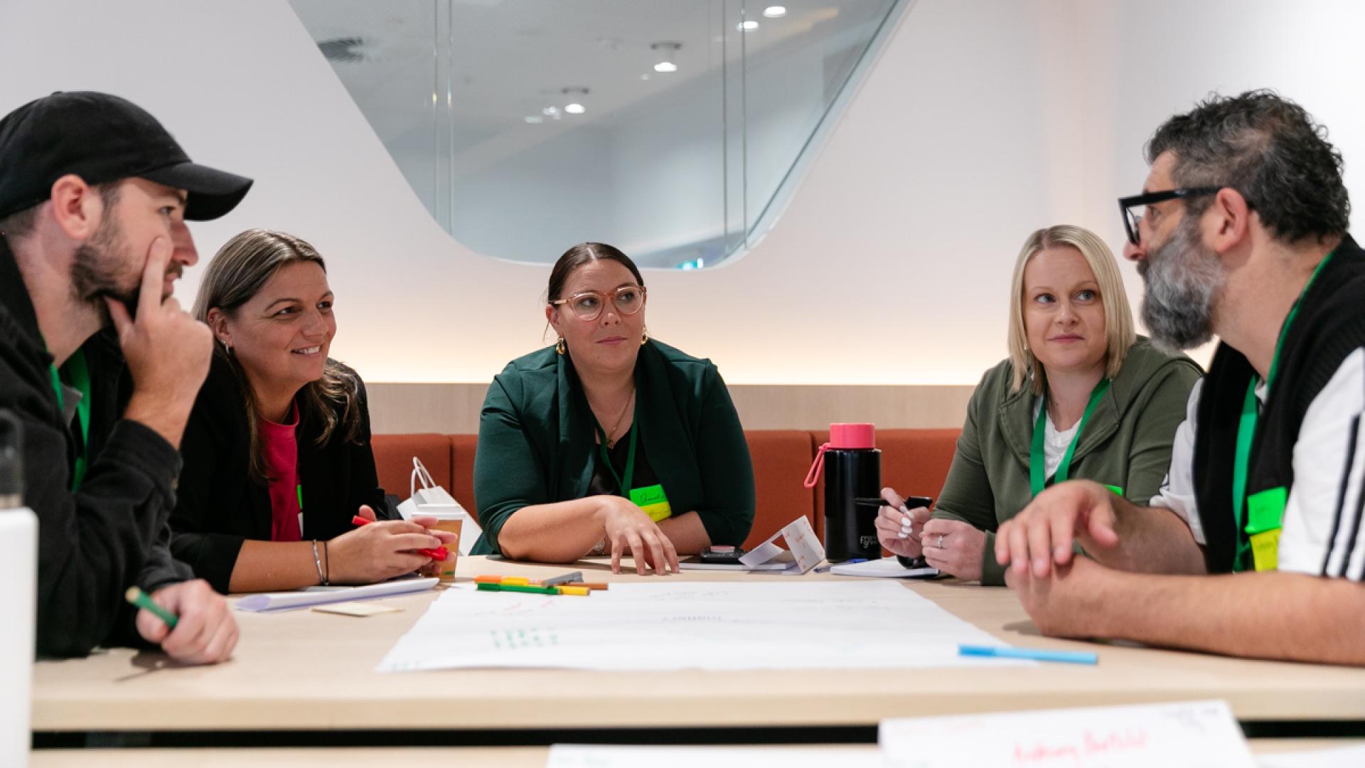 5 participants seated around table in discussion with paper and textas